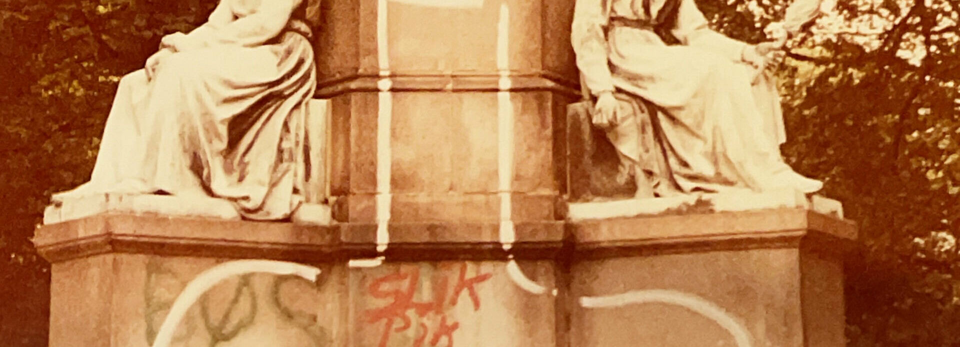 Foto av nederdelen av en skulpturgrupp med två sittande figurer. I mitten en grafitti-teckning av en penis.