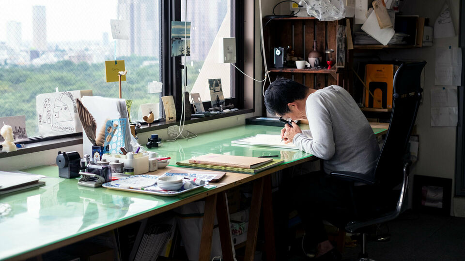En man sitter och ritar vid ett skrivbord som står framför ett fönster. Diverse ritverktyg på bordet och i taket hänger en samling skisser.