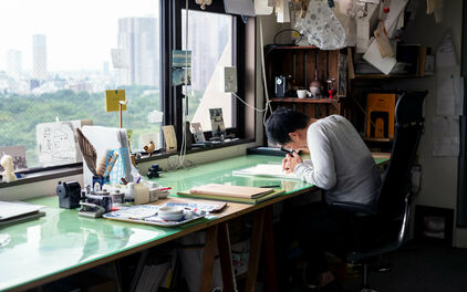 En man sitter och ritar vid ett skrivbord som står framför ett fönster. Diverse ritverktyg på bordet och i taket hänger en samling skisser.