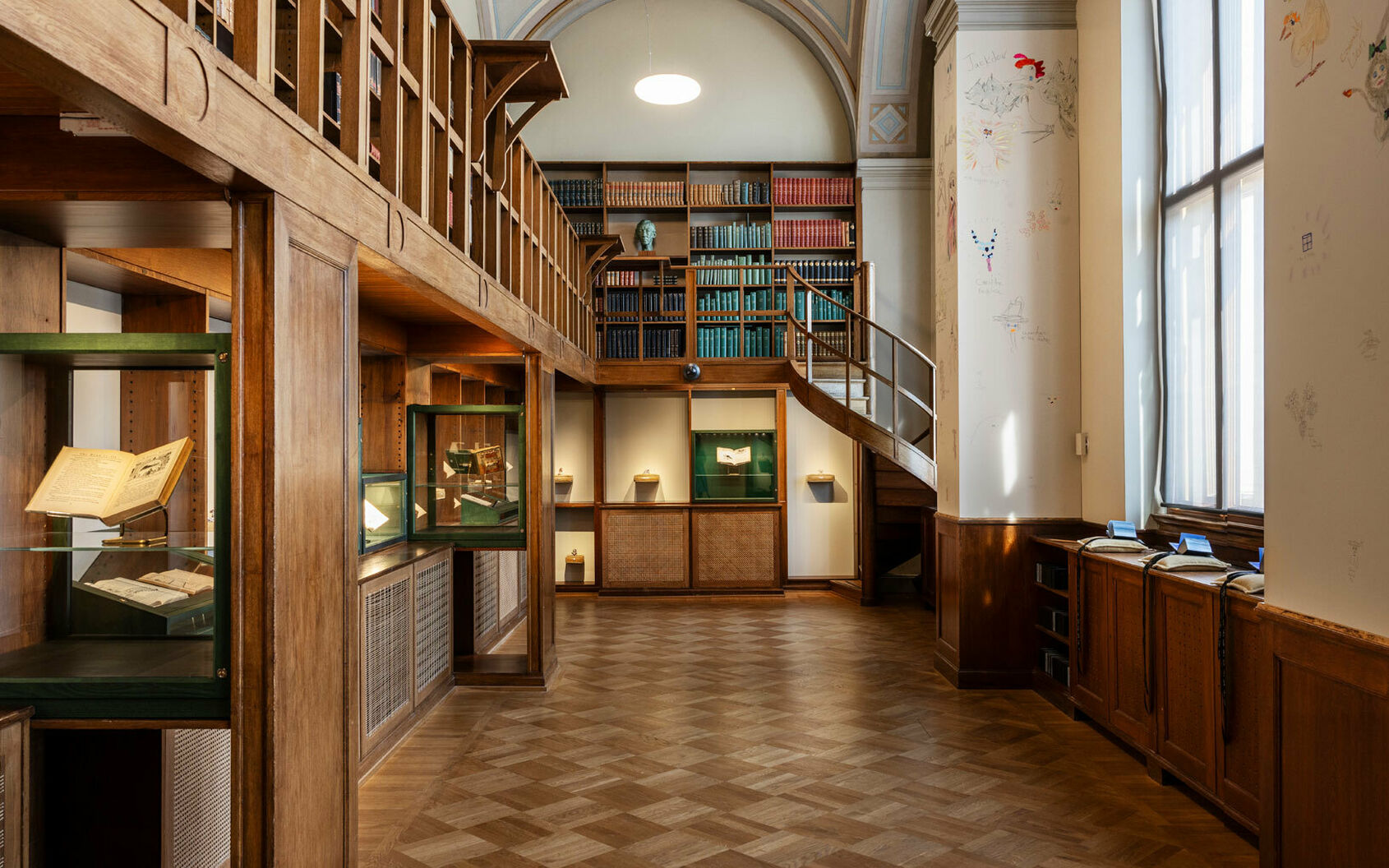 Interiör från ett gammalt bibliotek med böcker i träbokhyllor samt en konstinstallation i glasmontrar och med små böcker på kuddar i bibliotekets fönster.