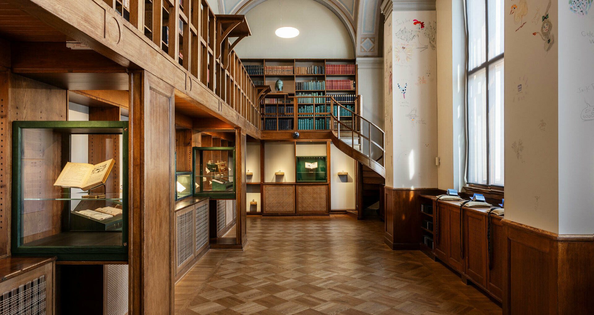 Interiör från ett gammalt bibliotek med böcker i träbokhyllor samt en konstinstallation i glasmontrar och med små böcker på kuddar i bibliotekets fönster.