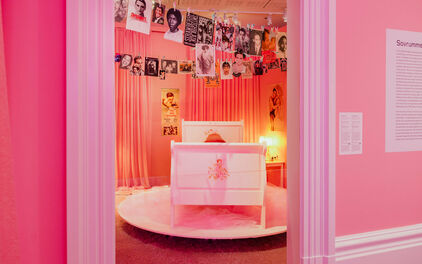 Foto från konstinstallation. En rosa säng i ett rosa rum. I taket linor med fästade bilder av olika personer.