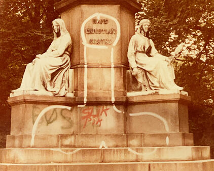 Foto av nederdelen av en skulpturgrupp med två sittande figurer. I mitten en grafitti-teckning av en penis.