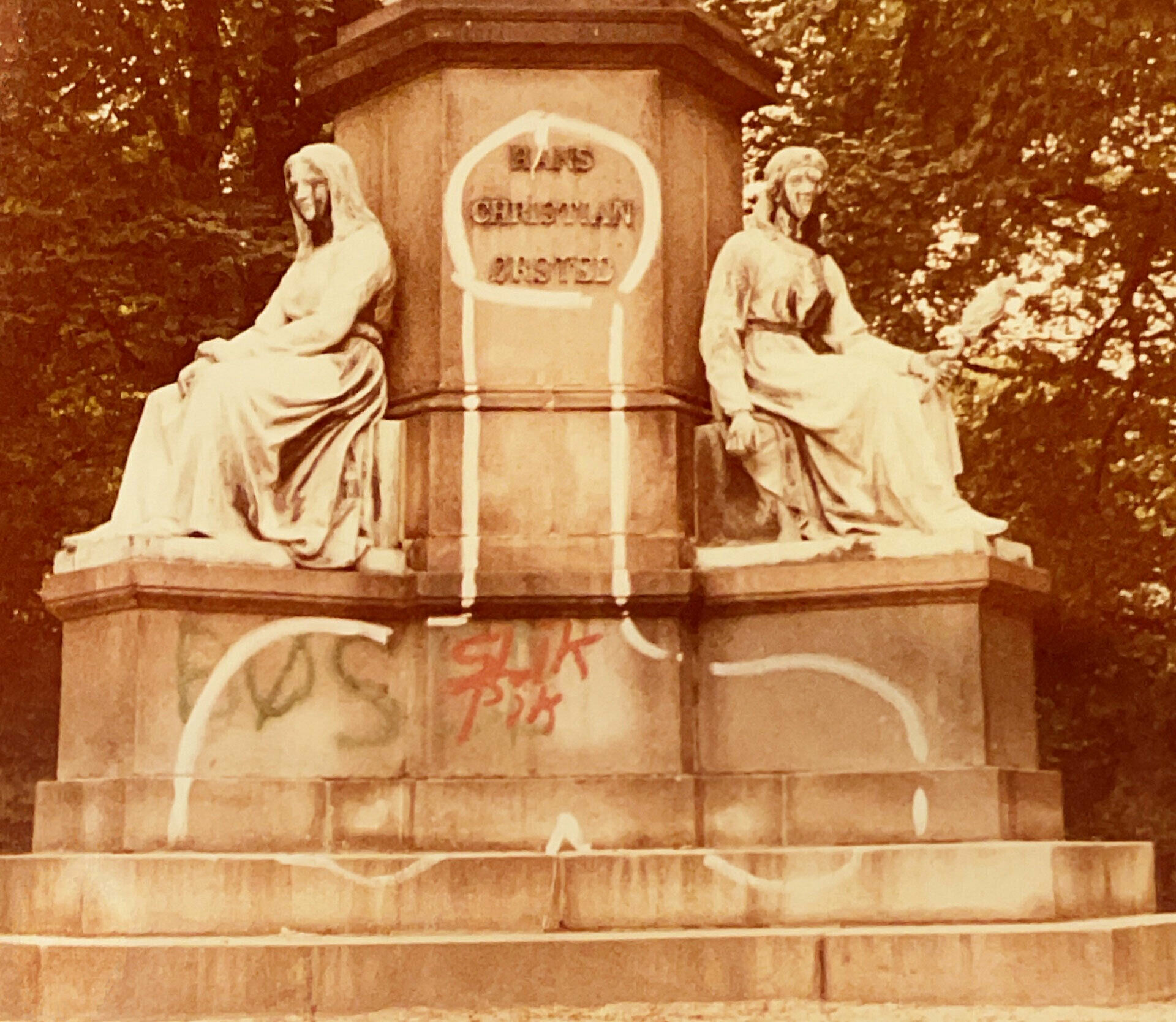 Foto av nederdelen av en skulpturgrupp med två sittande figurer. I mitten en grafitti-teckning av en penis.