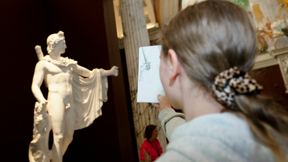 En flicka står framför en skulptur med ryggen mot betraktaren. Hon håller upp ett papper, där hon har ritat av en detalj av skulpturen, och jämför med förlagan.