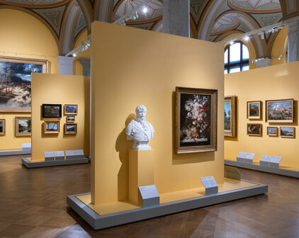 Foto av museisal med gula väggar. En skulpturbyst, föreställande en man, och målningar av landskap och blommor.