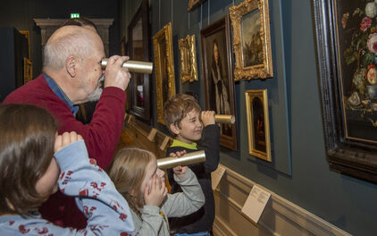En äldre man och tre barn tittar på tavlor i en museisal med hjälp av hemmagjorda kikare, eller tittinstrument.