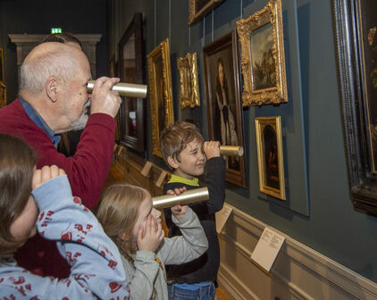 En äldre man och tre barn tittar på tavlor i en museisal med hjälp av hemmagjorda kikare, eller tittinstrument.