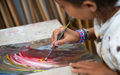 Ett barn som sitter vid ett bord i en ateljé och målar med akrylfärg i starka kulörer.